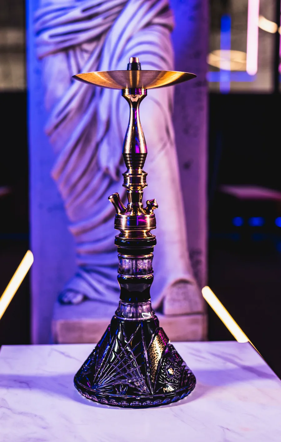 Cachimba MIG Tradi Lounge Edition con base de cristal tallado sobre una mesa blanca y neones rosas